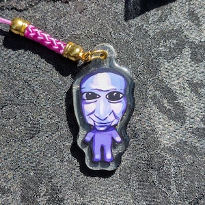 May include: A purple character charm with a large head and small body, featuring a smiling face with wide black eyes. The charm is made of clear acrylic with a glittery finish and is attached to a bright pink braided cord with gold metal hardware.
