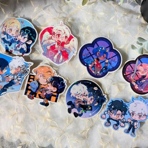 Op de afbeelding: Een verzameling kleurrijke stickers in anime-stijl met koppels in verschillende poses. De stickers tonen een reeks ontwerpen, waaronder personages met engelen vleugels en personages in een klaverblad vorm. Het kleurenpalet omvat blauw, roze en paars.