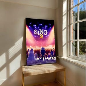 Op de afbeelding: Een ingelijste filmposter voor "Sing 2" met een podiumscène met silhouetten van personages. De titel "SING 2" wordt benadrukt door schijnwerpers. De poster wordt getoond op een bank met een geweven bovenkant en goudkleurige poten.