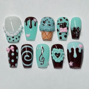 Puede incluir: Un conjunto de diez uñas postizas con varios diseños. Las uñas presentan una mezcla de colores, incluyendo verde azulado, marrón y blanco, con elementos decorativos como conos de helado, goteos de chocolate, lazos y notas musicales. Algunas uñas tienen lunares y acentos de corazón.