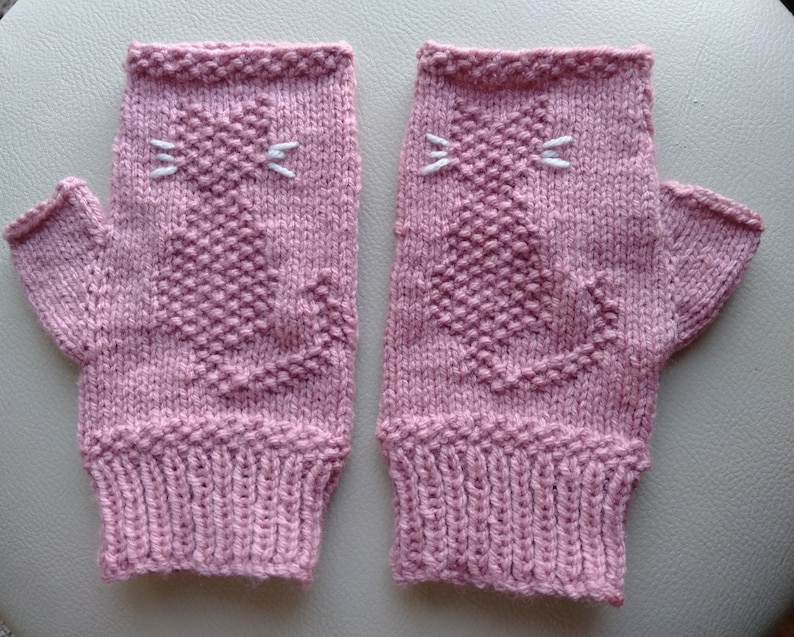 May include: A pair of pink knitted fingerless gloves with a white cat design. The cat has a white nose and whiskers.