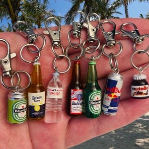 May include: A set of eight miniature beverage and food keychain charms, including Corona Extra, Evian, Budweiser, Heineken, Red Bull, Nutella, and a generic can of beer. The charms are attached to silver keychains with split rings.