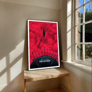 Puede incluir: Un póster enmarcado con un fondo rojo y una ilustración en blanco y negro. El póster presenta la palabra "HELLRAISER" y un diseño geométrico. El póster se exhibe en un banco tejido cerca de una ventana.