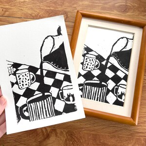 May include: Black and white linocut print featuring a checkered pattern with mugs. One print is framed in a wooden frame, and another is held in a hand. The mugs have different designs, including stripes and polka dots.