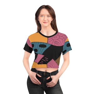May include: A black crop top featuring a colourful patchwork design. The design includes orange, pink, blue, and black patches with stitched details. The shirt has a black collar and short sleeves.
