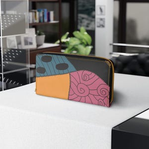 Peut inclure: Un portefeuille patchwork coloré avec une fermeture éclair dorée. Le design présente diverses formes et couleurs, notamment du bleu, de l'orange, du rose et du noir. La section rose présente un motif en spirale. Le portefeuille est posé sur une surface blanche.