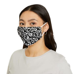 May include: A black and white face mask featuring a swirling, floral pattern. The mask has white ear loops and is worn by a person with dark hair and a light gray shirt. The mask covers the nose and mouth.