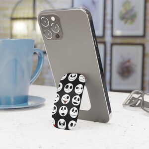 May include: A black phone grip featuring a pattern of white Jack Skellington faces from The Nightmare Before Christmas. The phone grip is attached to a silver smartphone, which is propped up on a white surface.