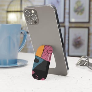 Peut inclure: Un téléphone portable avec un support de téléphone coloré sur le thème de Sally. La poignée présente un motif patchwork orange, rose, bleu et noir. Le téléphone est posé sur une surface blanche, avec une tasse bleue en arrière-plan.