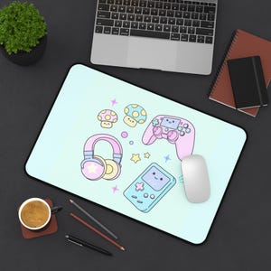 May include: A pastel blue mouse mat with a gaming theme. The design features a pink game controller, headphones, a handheld game console, and mushroom graphics. A white mouse sits on the mat, next to a laptop.