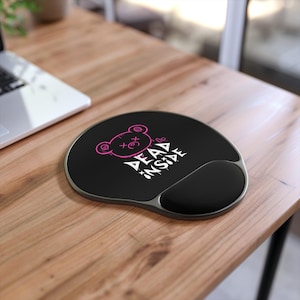 May include: Black ergonomic mousepad with a wrist rest. The mousepad features a pink graphic of a bear with the text "DEAD INSIDE" in white. The mousepad is on a wooden desk.