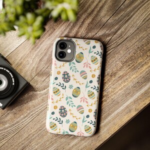 May include: A white iPhone case featuring a repeating pattern of Easter eggs, flowers, and leaves in pastel shades. The case is on a wooden surface, with a camera and greenery in the background.