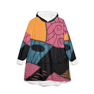 May include: A hooded blanket with a patchwork design in orange, pink, blue, and black. The blanket has a white fleece lining and cuffs. The design includes stitched details and a front pocket.