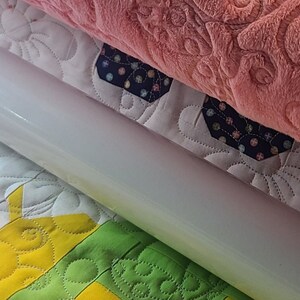 Peut inclure: Gros plan d'une courtepointe en cours de roulage. La courtepointe est faite de tissus de différentes couleurs, dont le rose, le violet, le blanc, le jaune et le vert. La courtepointe est matelassée avec un motif floral.