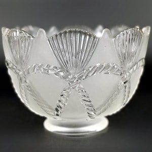 May include: A clear glass bowl with a frosted interior and intricate exterior detailing. The design features a rope-like pattern and fan-shaped accents, creating a textured and elegant appearance. The bowl sits on a small, round base.