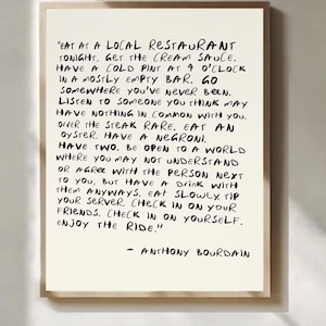 Puede incluir: Impresión enmarcada con texto manuscrito negro sobre fondo crema. El texto es una cita de Anthony Bourdain, que anima a disfrutar de la vida y a probar nuevas experiencias. El marco es de color marrón claro.