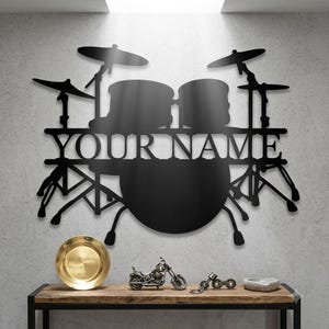 Op de afbeelding: Een zwart metalen wanddecoratie in de vorm van een drumstel met de tekst YOUR NAME in het midden geïntegreerd. Het ontwerp bevat bekkens, toms en het silhouet van een basdrum, wat een gedetailleerde weergave van een muziekinstrument vormt.