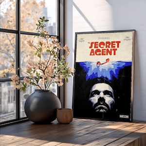 Puede incluir: Un póster enmarcado de la película "The Secret Agent" con un marco negro. El póster presenta la cara de un hombre, agua azul y texto rojo. Un jarrón negro con flores y una pequeña taza de madera están sobre una superficie de madera.