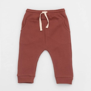 May include: Rust-colored baby pants with a waffle knit texture. The pants have a drawstring waist and two front pockets. The cuffs are ribbed. The pants are made of a soft, comfortable material.