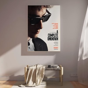 Könnte beinhalten: Ein gerahmtes Filmplakat für "A Complete Unknown" mit einem Mann mit Sonnenbrille. Das Plakat hängt an einer weißen Wand über einer kleinen Holzbank mit einer Decke und einer Tasse. Rechts hängt ein weißer Vorhang.
