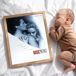 Może przedstawiać: Oprawiony plakat filmowy do filmu "Basic Instinct" z Michaelem Douglasem i Sharon Stone, z tytułem filmu w kolorze czerwonym. Śpiące dziecko w brązowym pajacyku leży obok oprawionego plakatu na białej powierzchni.