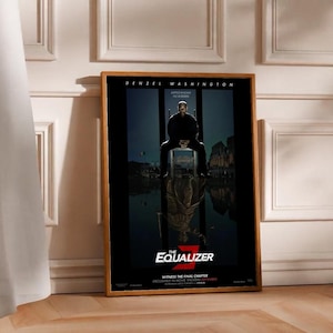 May include: A framed movie poster for "The Equalizer 3" featuring Denzel Washington. The poster has a dark color scheme with the title at the bottom. The poster is leaning against a wall with a wooden floor.