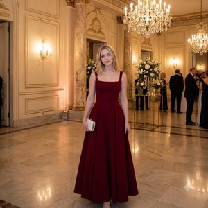 May include: A floor-length, sleeveless, deep burgundy gown with a square neckline and a fitted bodice that transitions into a full, flowing skirt. The dress is paired with a small, rectangular, off-white clutch held in one hand.