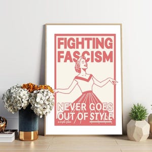 Puede incluir: Una impresión artística enmarcada con un diseño retro. La impresión presenta las palabras "FIGHTING FASCISM NEVER GOES OUT OF STYLE" en letras rojas. Una mujer con un vestido está ilustrada en rojo. La impresión se muestra sobre una superficie de madera.