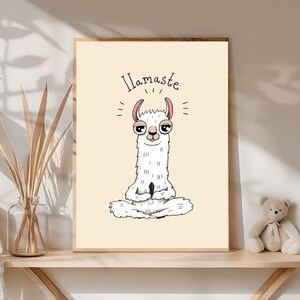 Op de afbeelding: Een ingelijste print met een cartoon lama in een yogahouding. De witte lama heeft roze oren en mediteert. De tekst "Llamaste" staat in handschrift boven de lama. De print heeft een lichtbeige achtergrond.