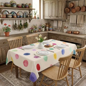 May include: A cream-colored tablecloth featuring a pattern of multicolored watercolor-style polka dots in shades of pink, blue, yellow, green, and purple, draped over a wooden dining table with four matching wooden chairs in a kitchen setting.