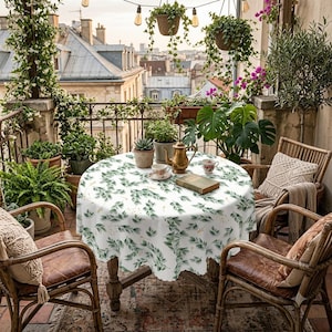 May include: A round table covered with a white tablecloth featuring a green leaf and gold accent pattern. On the table sits a gold teapot, two small floral teacups, and a closed book. The table is surrounded by four wooden chairs with cushions.