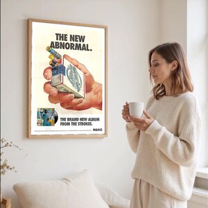 May include: A framed poster with the text "THE NEW ABNORMAL" and "THE BRAND NEW ALBUM FROM THE STROKES." The poster shows a hand holding a cigarette pack with colourful cigarettes. A woman in a cream sweater holds a white mug.
