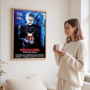 Puede incluir: Un póster enmarcado de la película "Hellraiser" con una imagen oscura de un personaje sosteniendo una caja de rompecabezas. El póster tiene el texto "Demon to some. Angel to others." y "He'll tear your soul apart."
