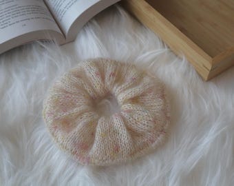 May include: A cream-colored, knitted scrunchie with subtle pink and yellow flecks. The hair accessory is circular and has a soft, textured appearance. The scrunchie is a hair accessory.