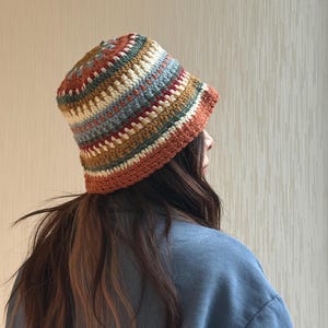 Peut inclure: Un chapeau seau crocheté à la main, aux couleurs vives. Le chapeau présente des rayures horizontales dans les tons orange, marron, crème, bleu et rouge. Le bord et la base du chapeau sont de couleur orange uni. Un accessoire de mode.