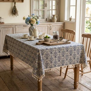 May include: A rectangular dining table covered with a cream-coloured tablecloth featuring a repeating blue damask pattern and a fringe trim. On the table sits a ceramic vase with flowers, a teapot, cups, a wooden cutting board, and a glass jar.