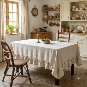 May include: A rectangular dining table covered with a solid light beige linen tablecloth featuring a decorative ruffled hem. The table is set with a ceramic bowl containing assorted fresh fruit and a single brass candlestick holder on top.