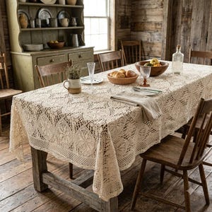 May include: A rectangular cream-coloured crochet tablecloth with an intricate floral and geometric pattern draped over a wooden dining table. The tablecloth features a scalloped edge and covers the surface, extending down the sides of the table.