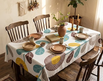 May include: A rectangular tablecloth featuring a white base with a repeating pattern of large stylised flowers and leaves in shades of teal, yellow, and brown. The fabric has a scalloped edge and is set on a wooden dining table with four chairs.