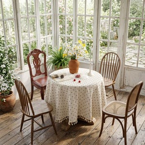 Peut inclure: Une table à manger ronde recouverte d'une nappe couleur crème à petits pois marron avec une bordure à franges. La table est entourée de quatre chaises en bois de styles différents et supporte un vase rempli de marguerites blanches.