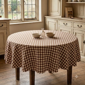 Peut inclure: Une table ronde recouverte d'une nappe à carreaux marron et blancs. Sur la table, il y a deux petits bols en céramique unis et une carafe en verre transparent. La nappe présente un bord festonné qui pend autour du périmètre de la table.
