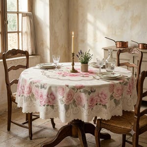 May include: A round tablecloth featuring a pattern of soft pink roses and green leaves on a cream background with a scalloped edge. The table is set with white plates, clear glass tumblers, a glass carafe, and a tall brass candle holder.