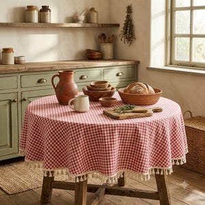 Könnte beinhalten: Ein runder Tisch mit einer rot-weiß karierten Tischdecke, die mit cremefarbenen Quasten verziert ist. Auf dem Tisch stehen ein brauner Keramikkrug, eine cremefarbene Tasse, gestapelte Holzschalen, ein geflochtener Korb mit Brot und ein Holzbrett.
