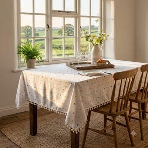 May include: A white tablecloth featuring a colourful polka dot pattern and a decorative pom-pom trim along the edges. The tablecloth is draped over a wooden table set with a wooden tray, two clear glasses, a white ceramic pitcher, and a small bowl.