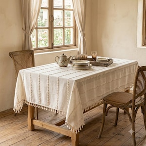 May include: A rectangular off-white tablecloth featuring vertical textured stripes and a decorative fringe of small tassels along the edges. The fabric has a woven appearance and covers a wooden dining table set with a ceramic teapot and plates.
