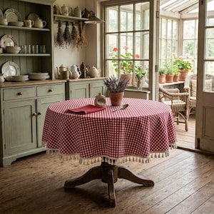 Può includere: Un tavolo rotondo in legno coperto da una tovaglia a quadretti rossi e bianchi con un bordo a frange decorative. Un piccolo vaso in terracotta contenente lavanda essiccata e un tovagliolo rosso piegato sono posti al centro del tavolo.