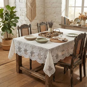 May include: A rectangular white lace tablecloth with a floral pattern and scalloped edges draped over a wooden dining table. The table is set with ceramic plates, a round woven placemat, a wooden tray with a ceramic pitcher, and a glass.