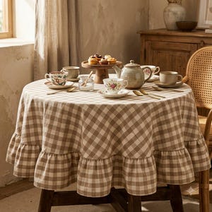 May include: A round table covered with a beige and white gingham tablecloth featuring a ruffled edge. The table is set with a ceramic teapot, several teacups and saucers with floral patterns, a small glass sugar bowl, and a wooden pedestal stand.