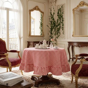 Peut inclure: Une table ronde à piédestal en bois recouverte d'une nappe à carreaux vichy rouge et blanc avec un ourlet à volants. Sur la table se trouvent une théière en porcelaine blanche, un pot à lait assorti, un petit vase en verre et une tasse.