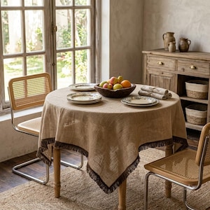 May include: A round table covered with a tan hessian tablecloth featuring dark brown fringe trim. The table is set with three white plates, a dark wooden bowl filled with assorted fresh fruit, and a stack of three folded linen napkins.